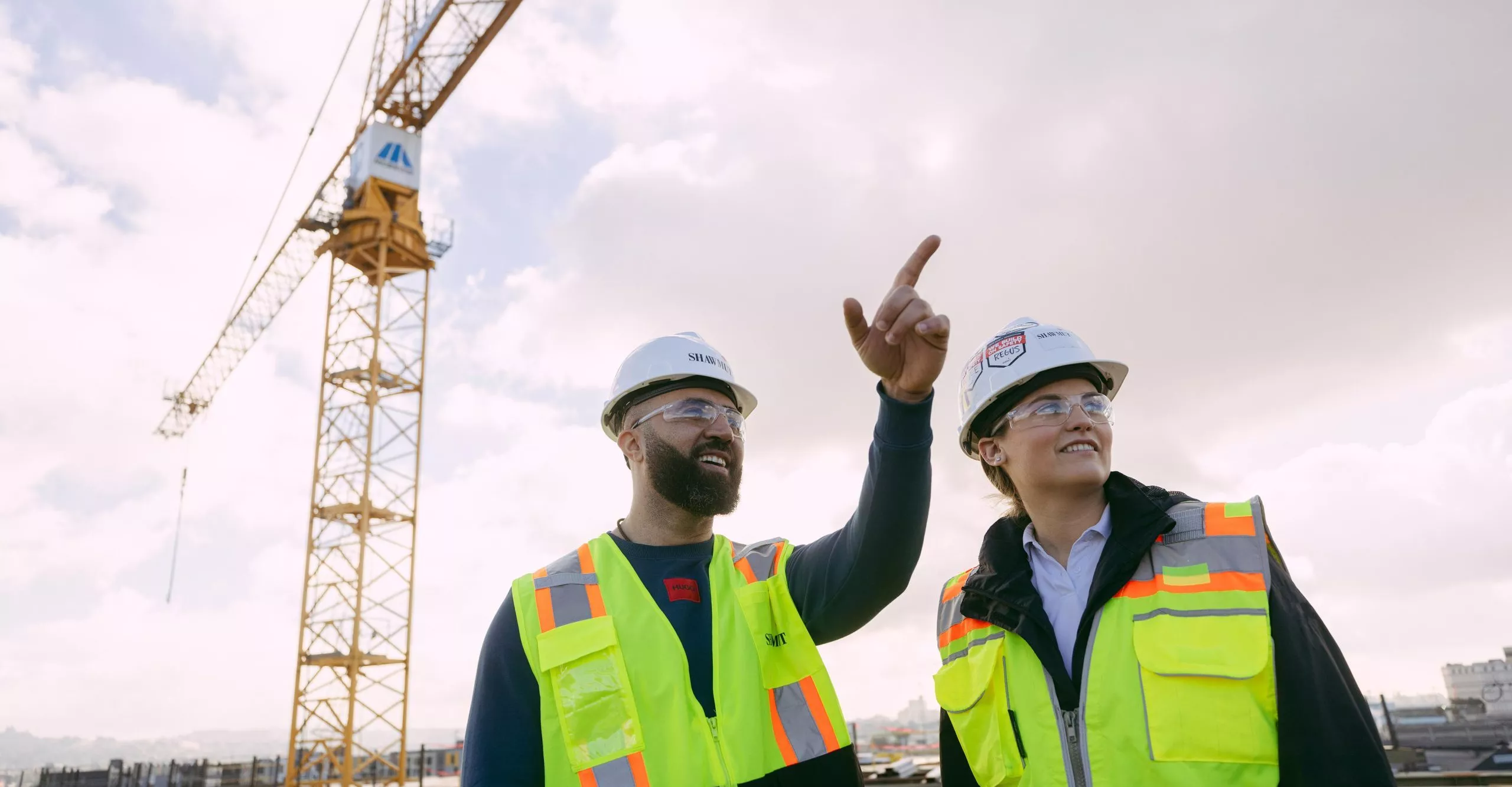 Two people in safety gear pointing into distance with crane in the background