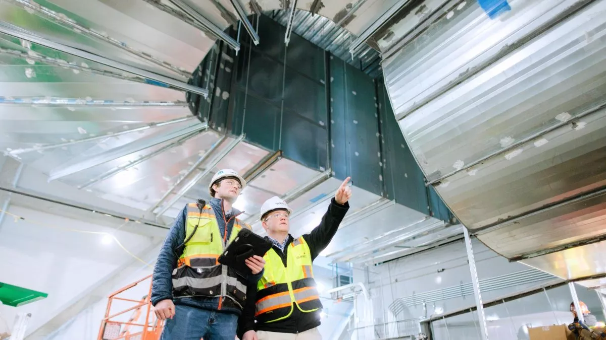 Two people in safety gear pointing to a vent in a building